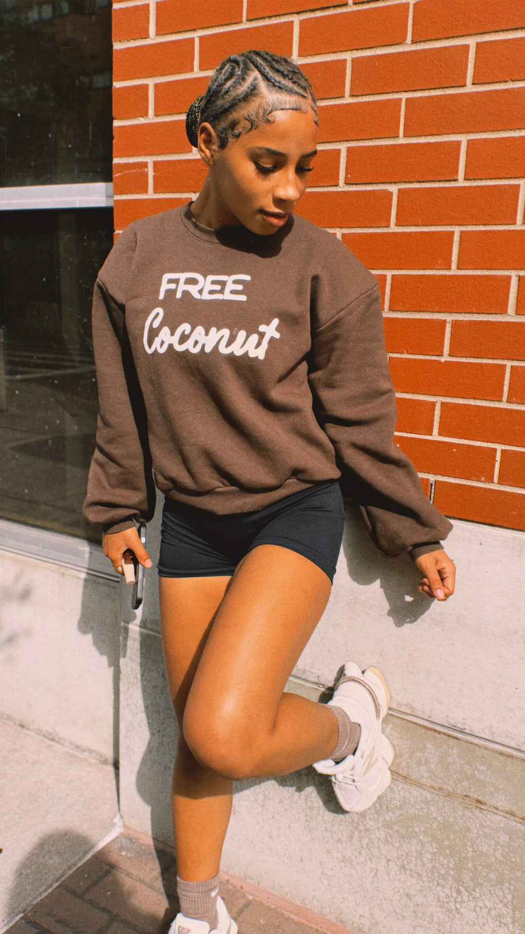 Person wearing a brown sweatshirt with 'Free Coconut' text, standing against a brick wall.