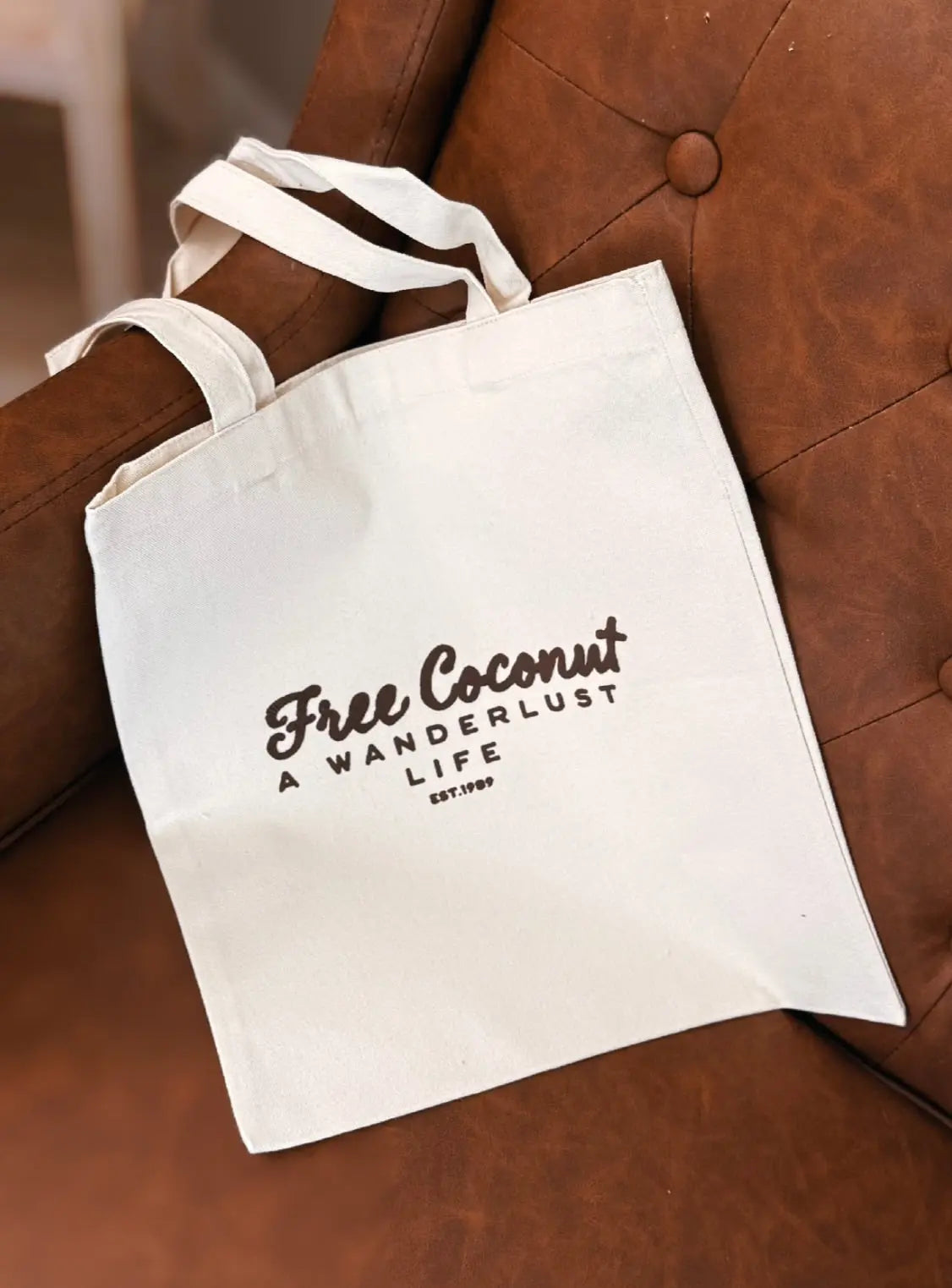 Cream canvas tote bag featuring the Free Coconut “A Wanderlust Life” logo in chocolate brown, styled on a brown leather chair. Minimal, aesthetic travel-inspired accessory.